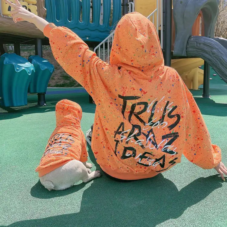 Parent and puppy wearing matching Orange (Autumn) hoodies with colorful splatter & text written on the back, twinning for a dog walk.